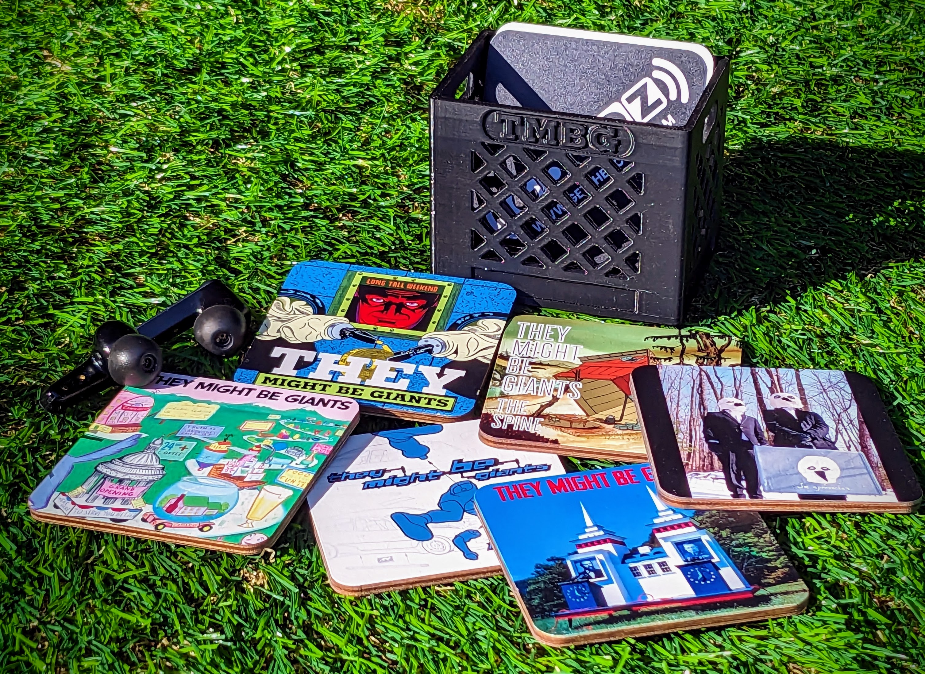 A wooden Custom Goodz card resting on grass near a crate of They Might Be Giants vinyl.