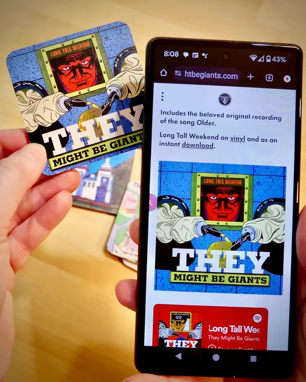 A They Might Be Giants 'Long Tall Weekend' Custom Goodz card next to a phone showing the band's website.