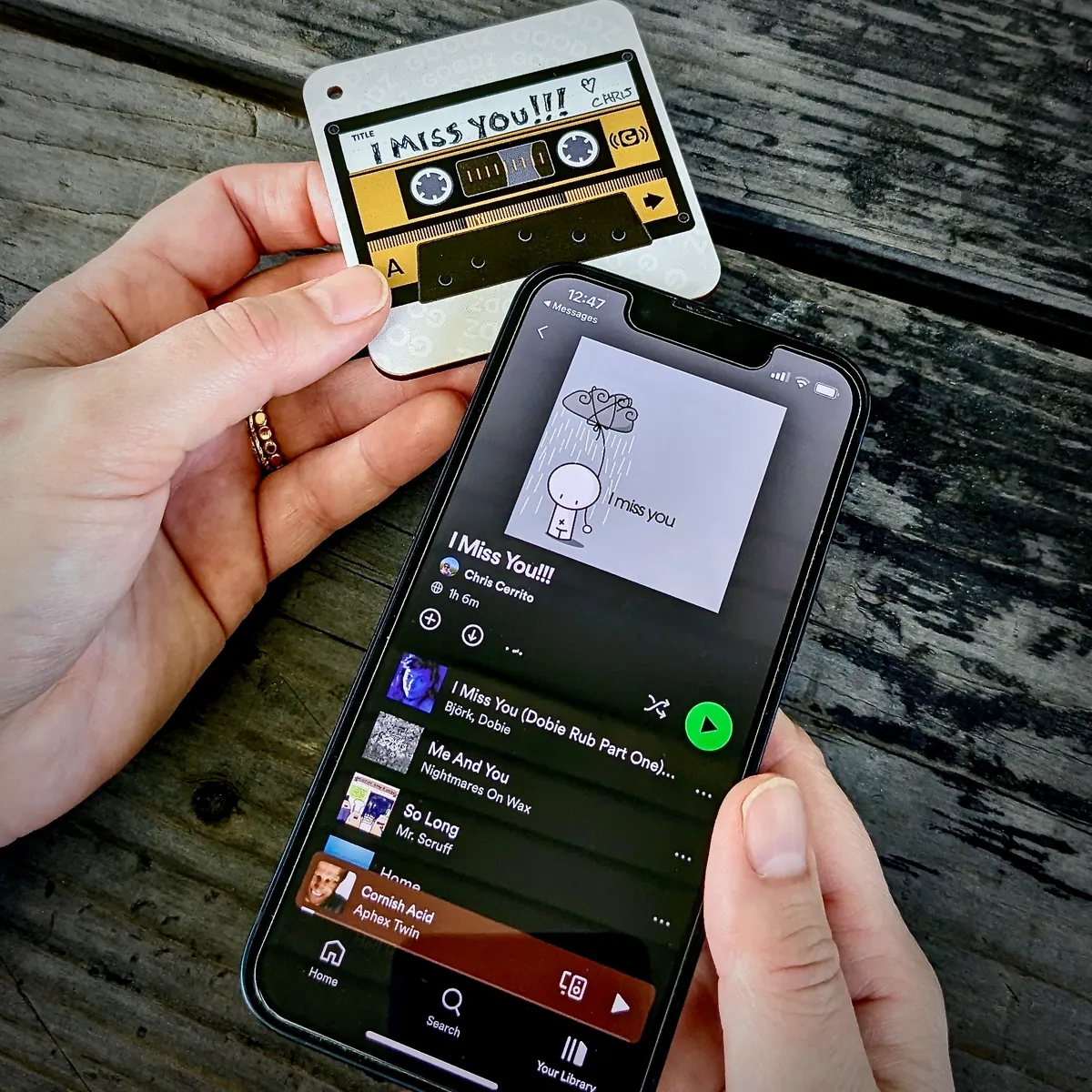 An 'I Miss You!!!' cassette-themed Mixtape Goodz held next to a phone showing the same-named Spotify playlist by Chris Cerrito.